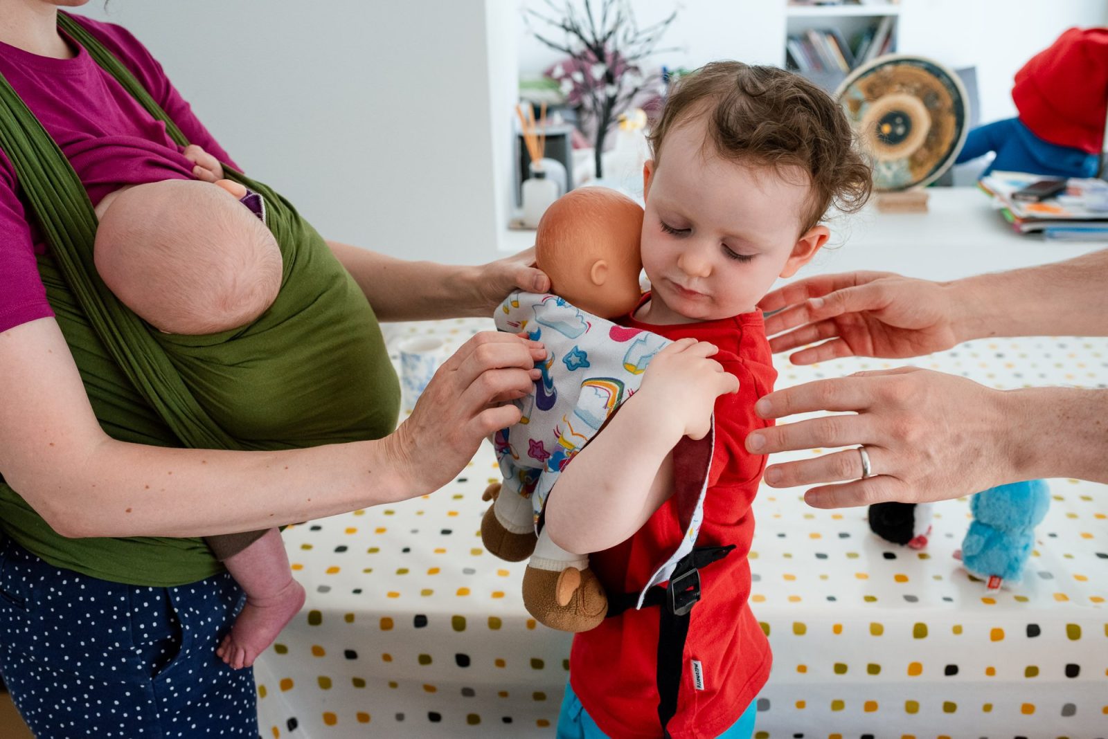 Toddler cuddling doll while mother holds baby in sling.
