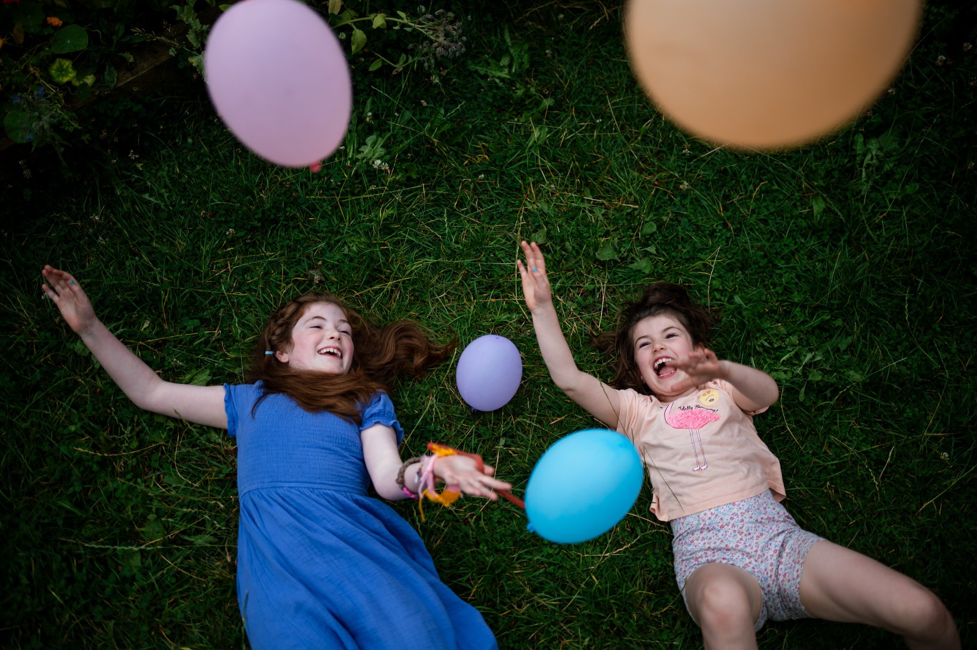 Two girls laughing with colourful balloons on grass.